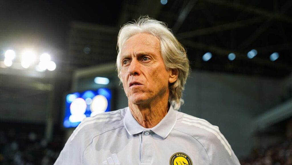 Former Al-Nassr Football Club manager Jorge Jesus looking sideways under floodlights in a stadium while wearing a white polo shirt with the yellow and blue club badge on it