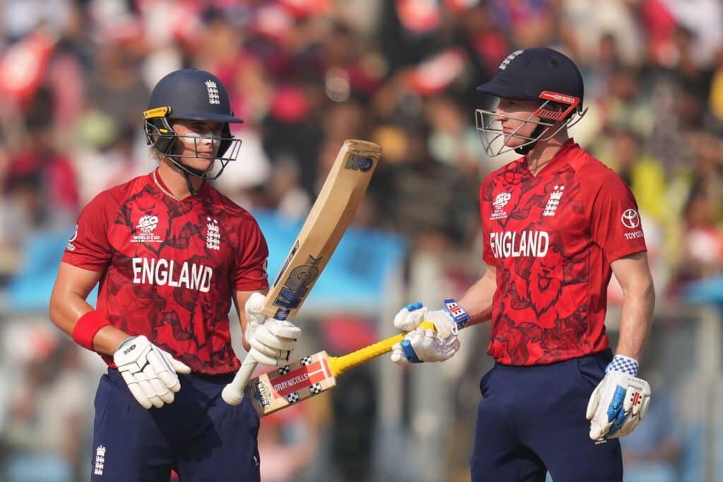 England T20 cricket players Jacob Bethell and Harry Brook celebrate during a game. The pair are wearing blue and white helmets and red shirts with England written on them, white gloves and blue trousers. Bethell is raising his bat
