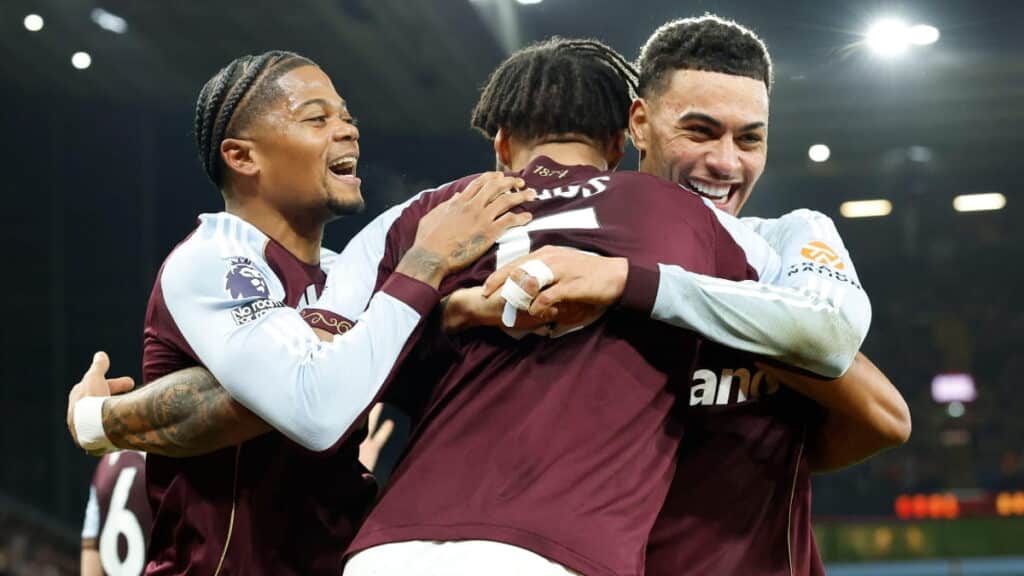 Aston Villa Football Club players celebrate a goal under floodlights in a stadium while embracing each other and wearing the club's home kit, with a maroon middle part of the shirt and light blue sleeves visible