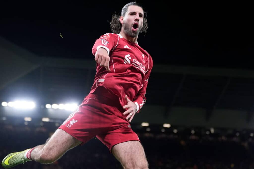 Liverpool Football Club footballer Dominik Szoboszlai jumps in the air to celebrate a goal in a dark stadium with shining floodlights in the background. The midfielder is wearing a predominantly red kit with white trim, has facial air and is punching with one arm