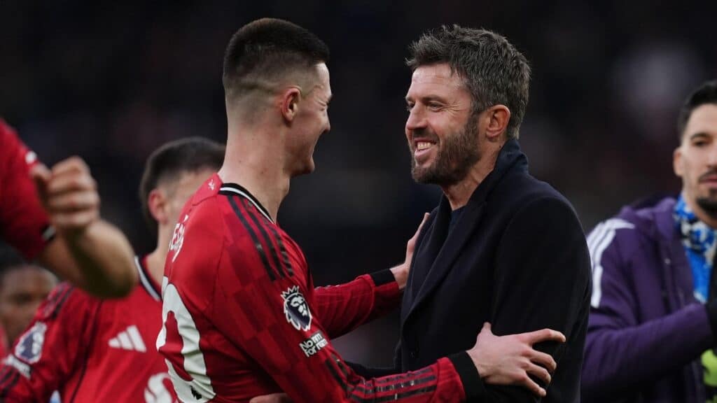 Manchester United Football Club manager Michael Carrick congratulates one of his players to his left, smiling and wearing a black top. Carrick has dark hair and a mustache and beard. His player is wearing a red, black and white Man United shirt, with other players appearing on the football pitch.
