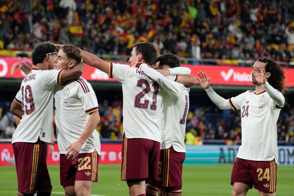 Mikel Oyarzabal celebrates after scoring for the Spain national team, embraced by his teammates with their backs to goals in white shirts and red shorts with their squad numbers showing
