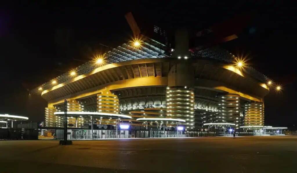O Stadio San Siro, sede dos clubes de futebol italianos da Serie A, Inter Milan e AC Milan, tem muitas luzes brilhando do lado de fora à noite, mas principalmente na escuridão e a entrada de concreto em primeiro plano.