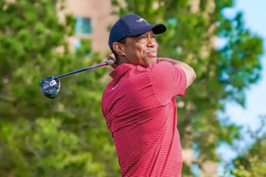 A photo of golfer Tiger Woods taking a shot on a golf course while wearing a light red shirt and wearing a dark blue baseball cap. Trees are visible blurred in the backrground