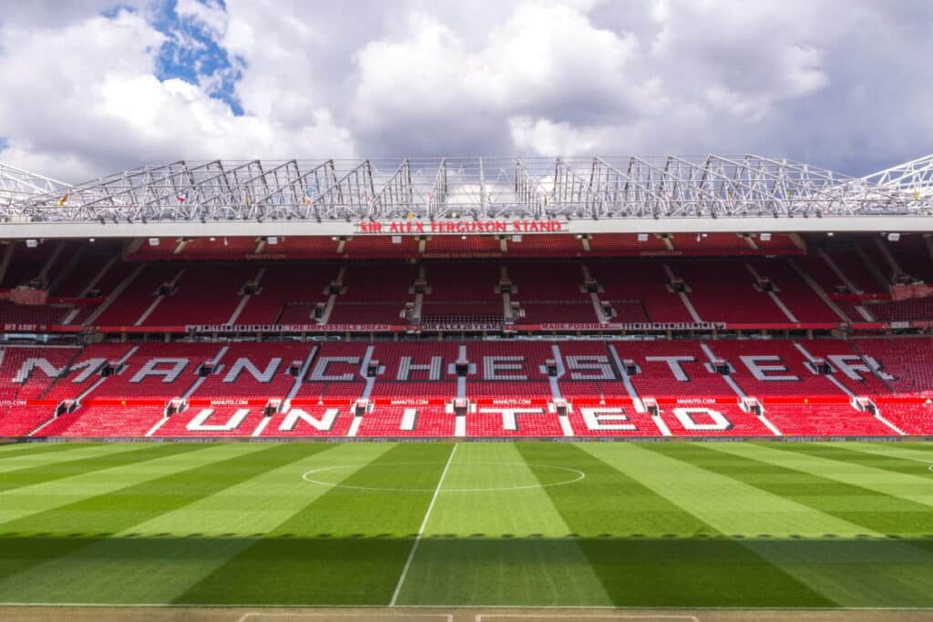 The Old Trafford stadium home of Manchester United Football Club showing a pitch in the foreground in two different shades of striped green leading to red and white seats spelling out the name of the club under a blue cloudy sky with shadows over part of the turf