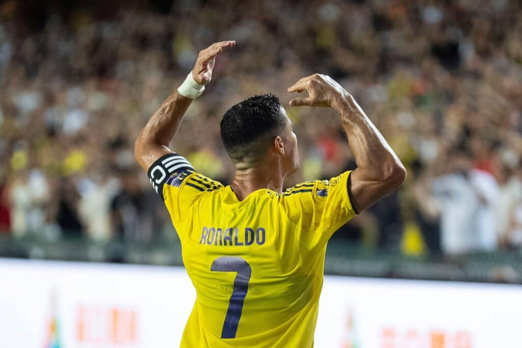 Footballer Cristiano Ronaldo playing football for Saudi Pro League club Al Nassr. Ronaldo is holding his arms in the air in a yellow shirt with a captain's armband and his name and number 7 number in blue, as well as blue trim. A blurry crowd is visible