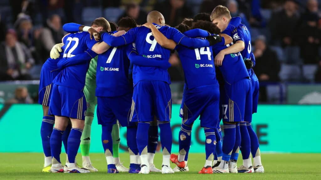 Leicester City Football Club footballers form a circular huddle of players on a green pitch. The players are linking arms and wearing blue shirts with white numbers on the back and blue shorts and socks with yellow trim, as well as different coloured boots, some of which are white, blue and red with black details. A crowd peppered with some empty seats is blurrily visible in the background
