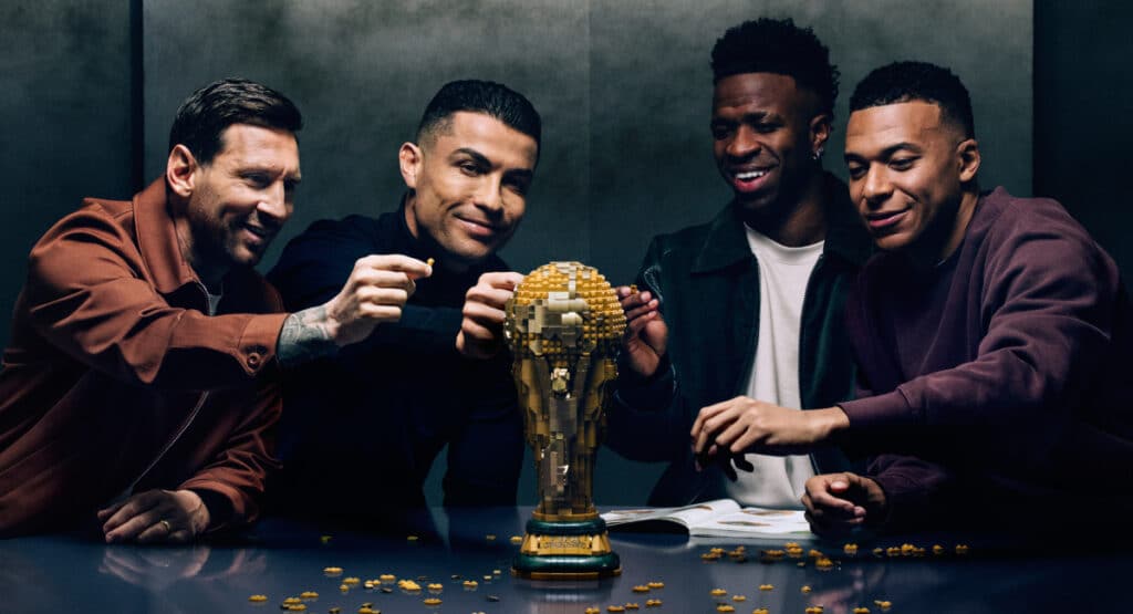 Foto dos jogadores de futebol Lionel Messi, Cristiano Ronaldo, Vinicius Jr e Kylian Mbappe sentados ao redor de uma mesa sorrindo para um modelo Lego da Copa do Mundo FIFA. Messi, Ronaldo e Mbale levantam cada um a mão em direção ao troféu e usam blusas escuras. Vinicius Jr. usa top preto e camiseta branca