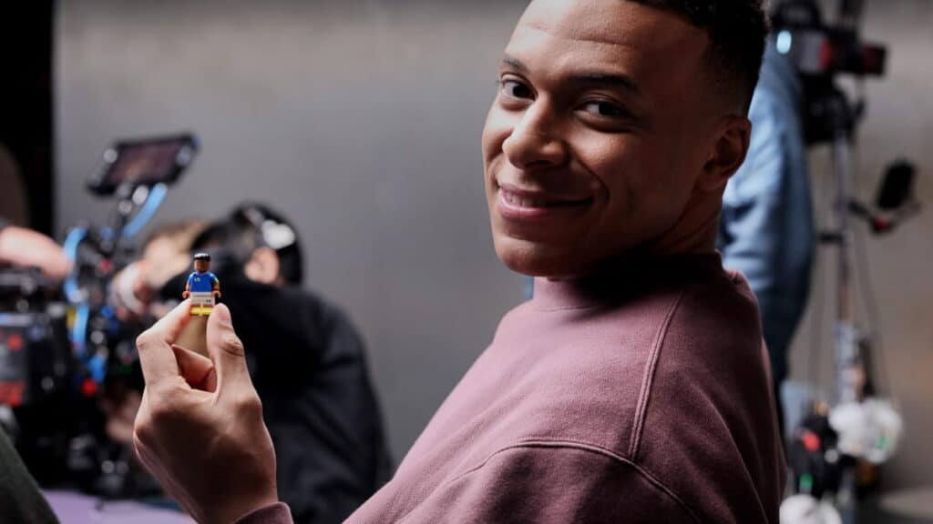 Footballer Kylian Mbappe grinning while a filming camera is visible in the background. Mbappe is wearing a dark-coloured top