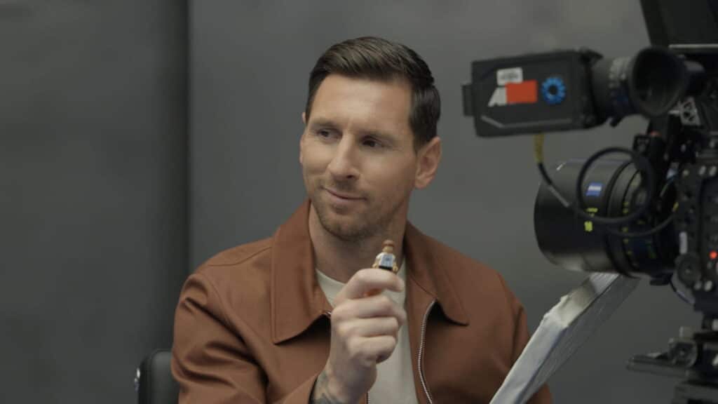 Football player Lionel Messi sitting in the room while being filmed. The camera is seen from the front and messi is wearing a brown jacket and white t-shirt while raising his hand.