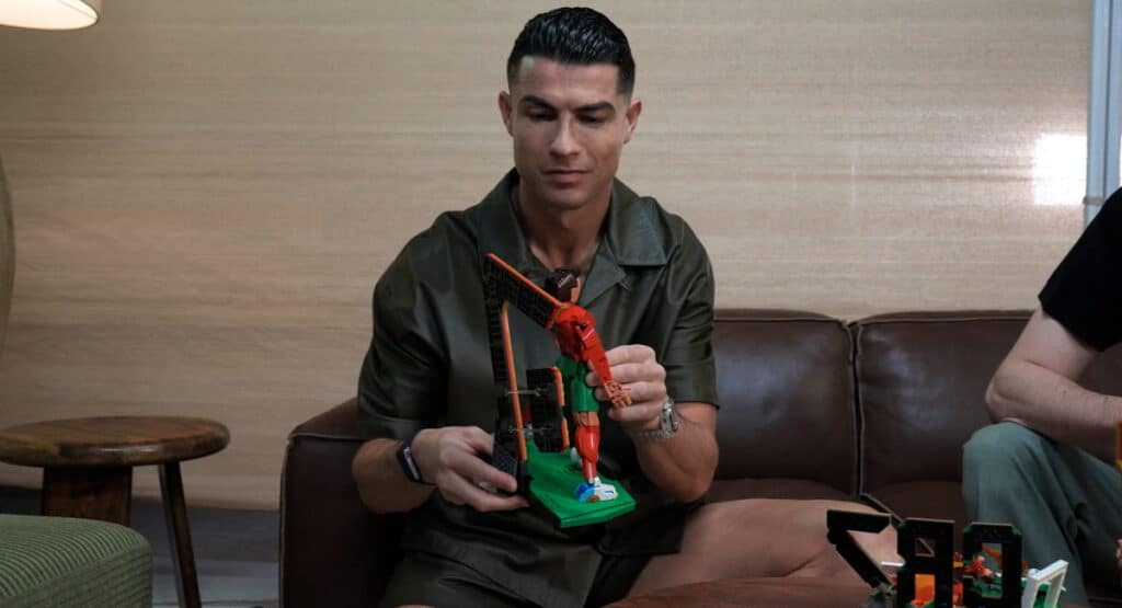 Footballer Cristiano Ronaldo sitting on a sofa playing with a Lego toy on a table. Ronaldo is wearing a dark top and shorts