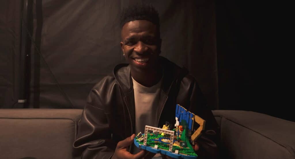 Soccer player Vinicius Junior laughs while holding up a lego toy in a dark room. Vinicius Junior is wearing a dark colored jacket
