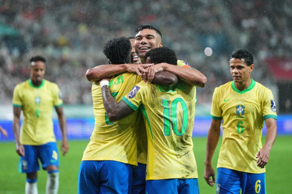 O jogador de futebol Casemiro comemora um gol enquanto joga pela Seleção Brasileira de Futebol. O jogador de futebol vira-se para a câmera e junta os braços para um abraço, enquanto Casemiro olha para a direita, boquiaberto e sorridente. Os jogadores estão no estádio e vestem camisas amarelas com numeração e letras verdes nas costas