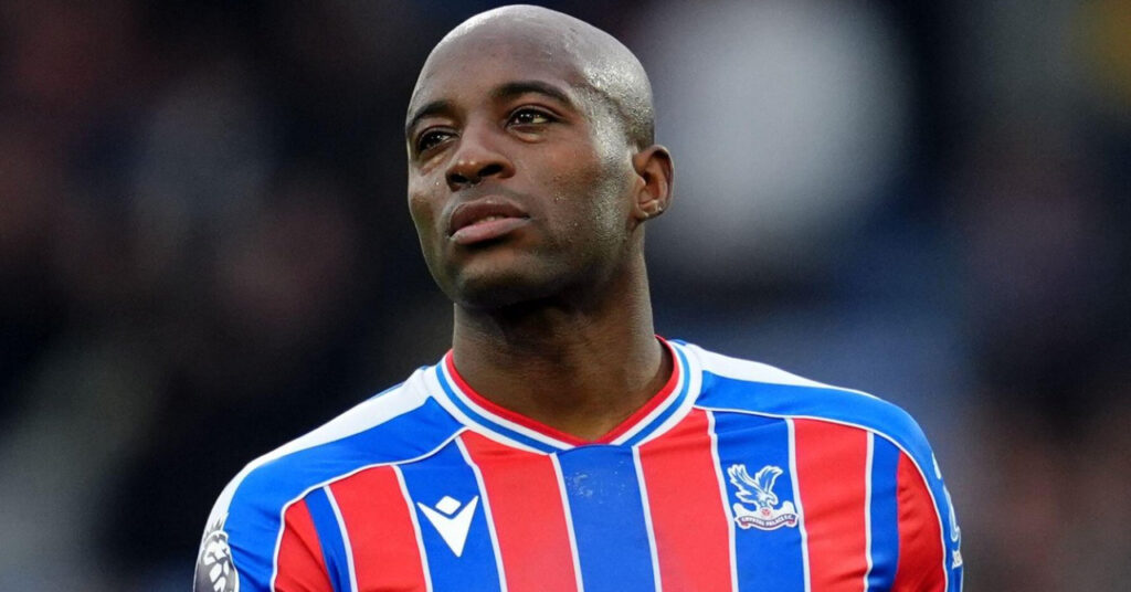 Crystal Palace footballer Jean-Philippe Mateta playing football for the club while looking to his right on a pitch in a red and blue striped shirt with white trim. The Palace Football Club badge is to the left of his chest, with a kit logo in white to the left
