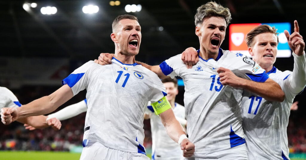 Footballer Edin Dzeko celebrates scoring for Bosnia and Herzegovina national football team. The striker has his arms outstretched and mouth open in a stadium under bright lights and is being embraced by two teammates to his left and one to his right as he looks at the crowd. The players are wearing white shirts with blue trim and blue numbers on their chests