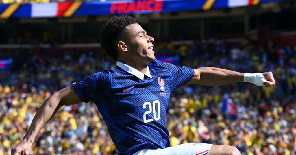 A jogadora de futebol francesa Desiree Du comemora o gol da França saltando no ar em um estádio lotado com grama verde. O jogador de futebol veste camisa azul com número 20 branco, calção branco, meias vermelhas e chuteiras pretas e salta com a perna direita na frente da esquerda e os braços estendidos. A palavra França pode ser vista em placas ao redor das arquibancadas