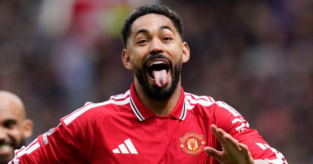 Footballer Matheus Cunha of Manchester United Football Club celebrates scoring their second goal during the Manchester United vs Nottingham Forest Premier League match at Old Trafford, Manchester. The player is wearing a red shirt with white trim and white snapdragon logo and Adidas logo and the club crest on his chest, with white shorts with black trim. Cunha has his tongue out and is looking ahead with a beard and moustache. A crowd is blurrily visible in the background, as is teammate Bryan Mbeumo, who is wearing the same kit