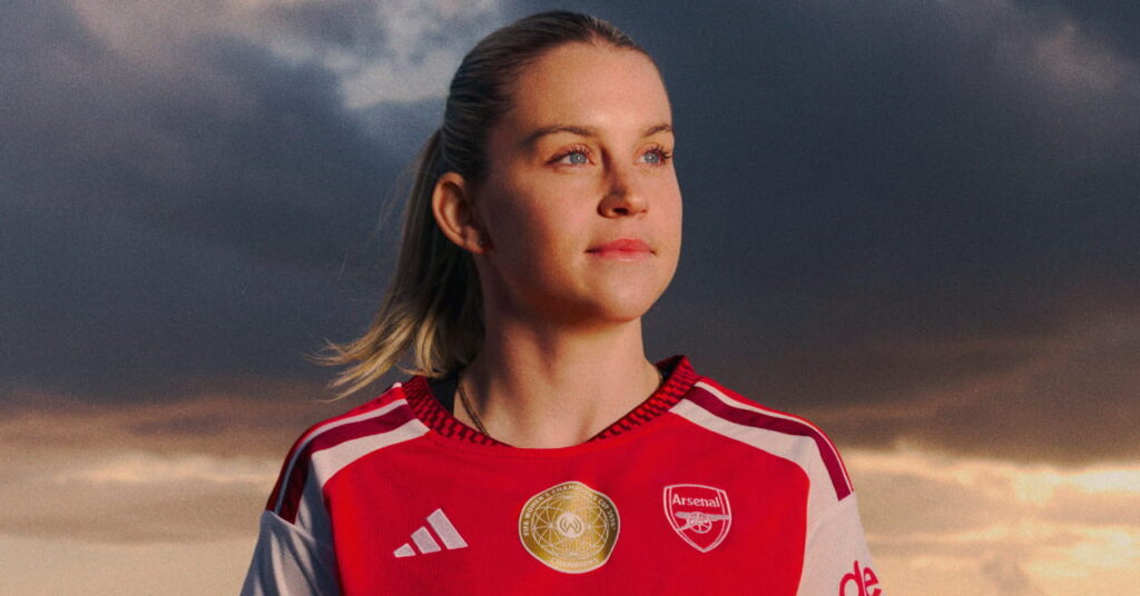 Arsenal football player Alessia Russo modelling the new club home shirt for the 2026/27 season, which is predominantly red with darker red trim and white details. The club crest and Adidas logo are in white either side of the chest with a gold circle in the middle. Russo is looking to her left under an atmospheric sky outside
