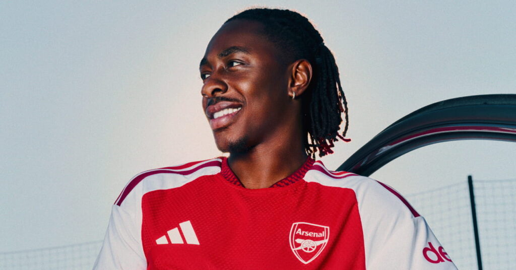 Arsenal football player Eberechi Eze modelling the new club home shirt for the 2026/27 season, which is predominantly red with darker red trim and white details. The club crest and Adidas logo are in white either side of the chest with a gold circle in the middle. Eze is looking to his right in an outdoor setting while leaning on a car door and smiling