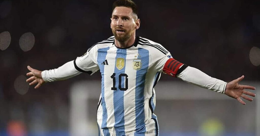 Footballer Lionel messi looking to his right with his arms outstretched while playing for the Argentina national football team. Messi is wearing a blue and white striped shirt with the number 10 in black on his chest, white forearm sleeves and a red captain's armband. A stadium background is blurrily visible