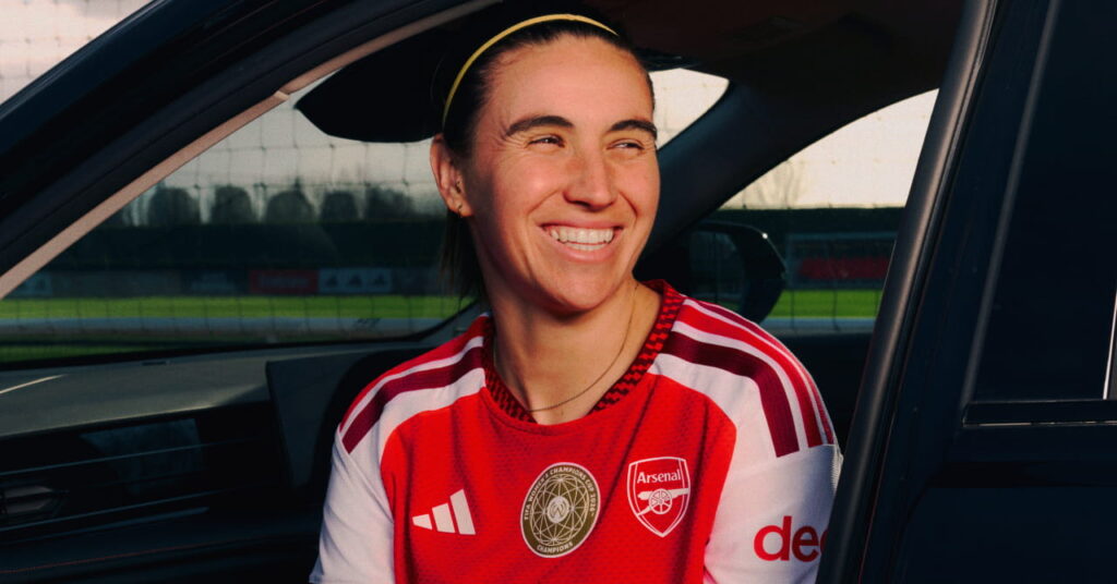 Arsenal football player Mariona Caldentey modelling the new club home shirt for the 2026/27 season, which is predominantly red with darker red trim and white details. The club crest and Adidas logo are in white either side of the chest with a gold circle in the middle. Caldentey is looking to her left while sitting in a car outside a training ground pitch smiling