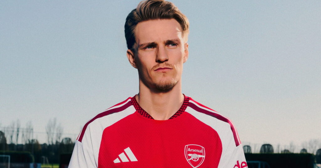 Arsenal football player Martin Odegaard modelling the new club home shirt for the 2026/27 season, which is predominantly red with darker red trim and white details. The club crest and Adidas logo are in white either side of the chest with a gold circle in the middle. Odegaard is looking to his left while standing under a blue sky