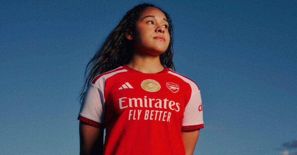 Arsenal football player Olivia Smith modelling the new club home shirt for the 2026/27 season, which is predominantly red with darker red trim and white details. The club crest and Adidas logo are in white either side of the chest with a gold circle in the middle. Smith is looking to her left while standing under a blue sky