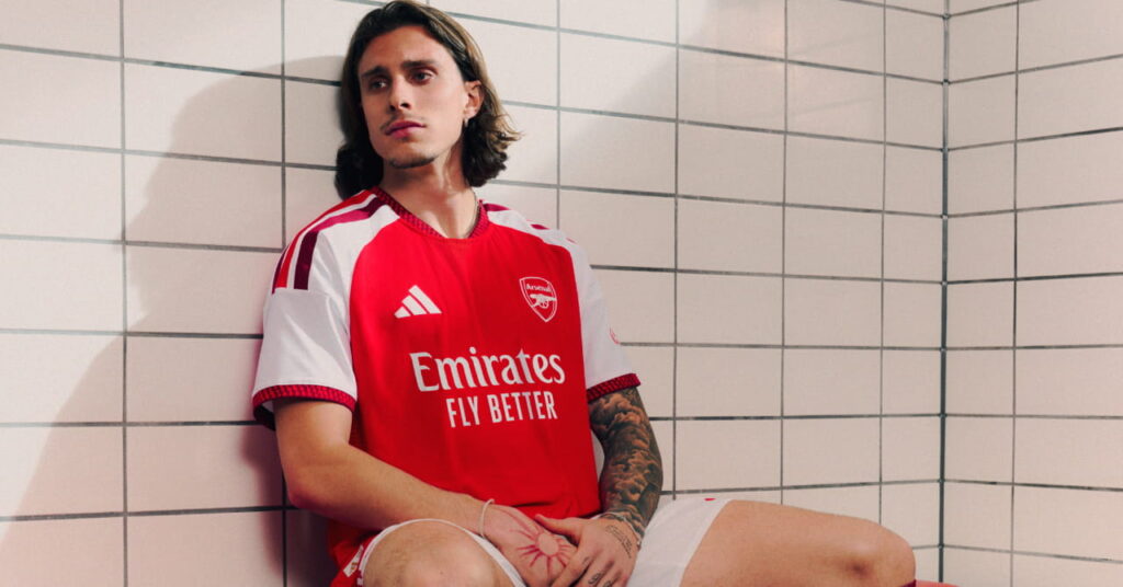 Arsenal football player Riccardo Calafiori modelling the new club home shirt for the 2026/27 season, which is predominantly red with darker red trim and white details. The club crest and Adidas logo are in white either side of the chest. Calafiori is sitting on a bench in a changing room looking to his right against a tiled white wall