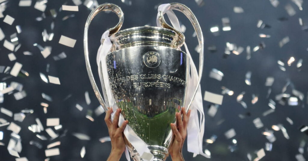 Um grande plano de um troféu de prata para o jogo de futebol da UEFA Champions League, um par de mãos entrelaçadas sob uma forma oval e segurando-o para o céu, com uma fita prateada enrolada em torno dele.