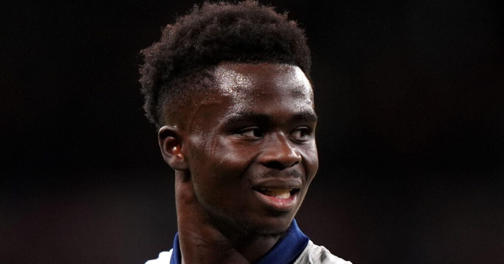 Footballer Bukayo Saka looking to his left with each arm out and an open mouth while playing for the England national football team. Saka is wearing a white shirt with blue trim and the number 7, Nike manufacturer tick and England crest on his chest against a dark outdoor backdrop
