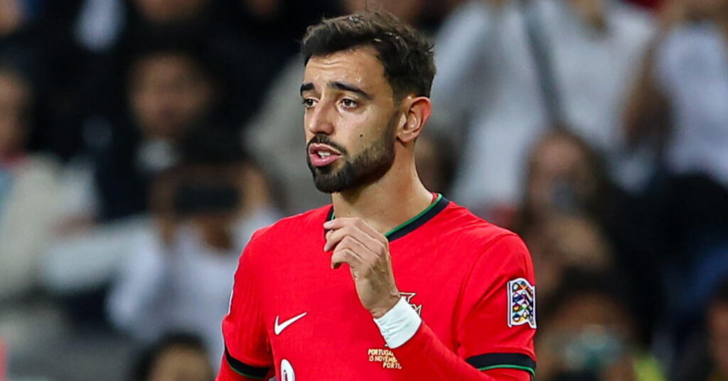 A photo of footballer Bruno Fernandes looking to his right while on a football pitch. The football player is wearing a Portugal national football team kit in red with green and black trim and a white nike logo and number on his chest. A crowd is blurrily visible in the background in the stadium
