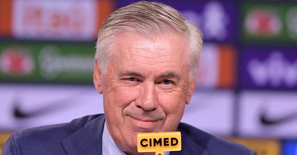 Uma foto do técnico de futebol Carlo Ancelotti falando ao microfone. O técnico do Brasil sorri e veste uma jaqueta azul escura e gravata combinando com uma camisa branca com o logotipo do patrocinador levemente visível atrás dele.