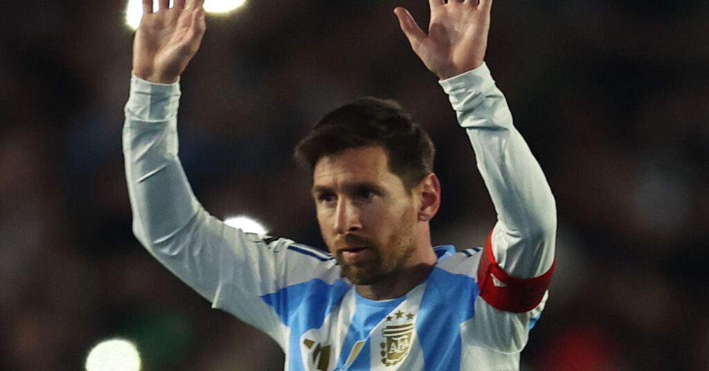 Foto do jogador de futebol Lionel Messi em campo olhando para a direita e levantando os braços Messi veste a camisa branca da seleção argentina de futebol, com o distintivo nacional dourado no peito e a braçadeira de capitão vermelha.