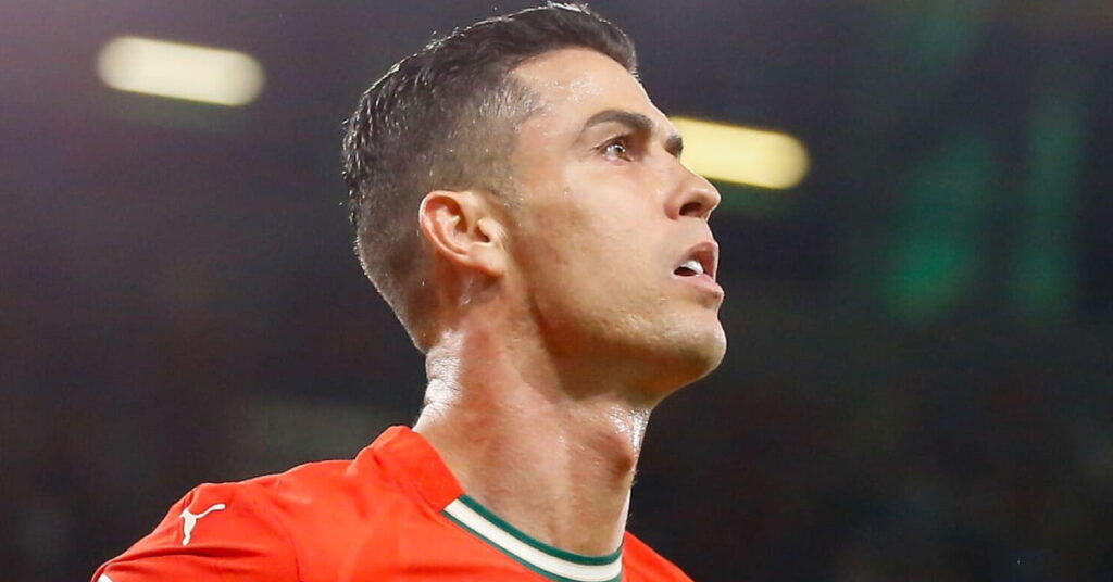 A photo of footballer Cristiano Ronaldo looking to his right while on a football pitch. Ronaldo is wearing a Portugal national football team kit in red with green and black trim and a white nike logo and number on his chest. Lights are blurrily visible in the background in the stadium