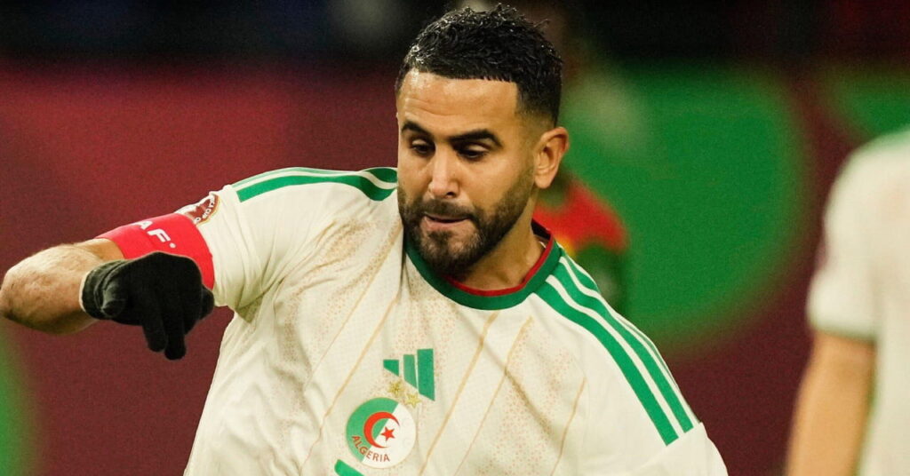 A photo of footballer Riyad Mahrez taking a penalty while playing for the Algeria national football team on a grass pitch. Mahrez is wearing black gloves and a white kit with green trim, a green number 7, Algeria badge and Adidas logo on his chest and blue boots. Other players are blurrily visible in the background