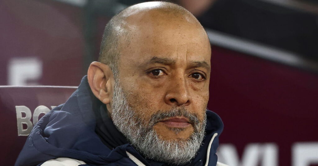West Ham United Football Club manager Nuno Espirito Santo looking to his left while sitting in a chair in a technical area at the team's London Stadium Home. Santo has a white and grey beard and moustache and is wearing a dark club jacket with white details of manufacturer Umbro and the club crest on his chest