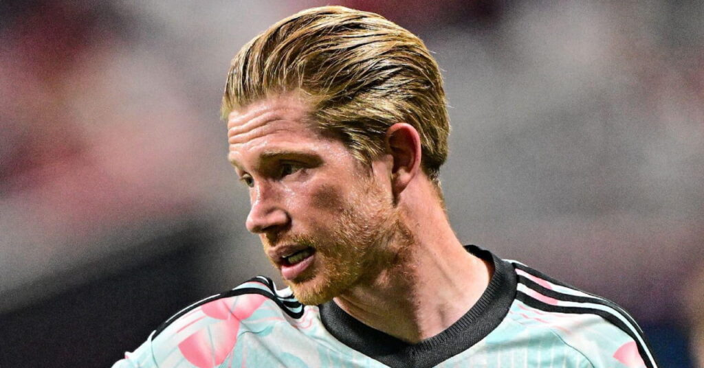 A photo of footballer Kevin De Bruyne looking to his right while standing on a pitch smiling and playing for the Belgium national football team in a light blue and pink shirt with a black collar and black lines running down the shoulders