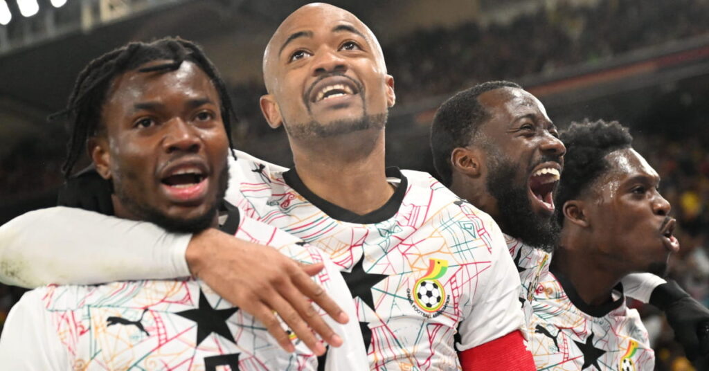 Jogadores de futebol ganenses comemoram um gol enquanto jogam pela seleção nacional de futebol em um estádio vestindo uma camisa branca com detalhes pretos e uma estrela preta no centro do peito.