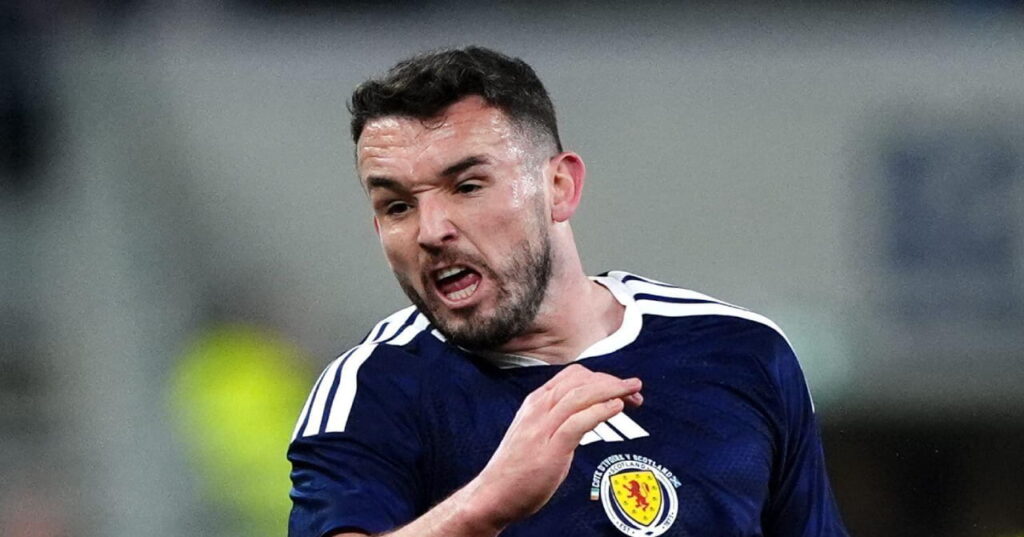 O jogador de futebol John McGinn vestindo uma camisa de futebol da seleção escocesa com um top azul escuro com uma faixa branca da adidas no ombro e uma bandeira nacional amarela no peito durante um jogo no estádio. McGinn olha para a direita