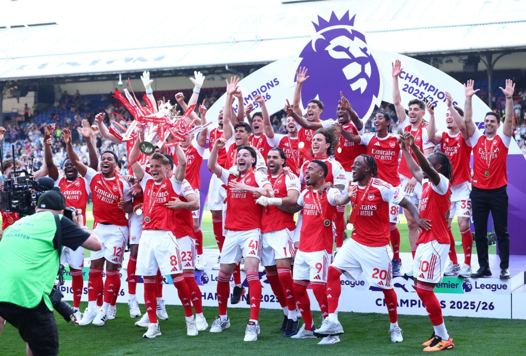 A photo of Arsenal footballers celebrating with the Premier League trophy on a pitch in front of a white competition stand showing the logo of a lion in blue above them. The group are standing on a grass pitch wearing red and white shirts, white shorts with red trim and red socks. Jurrien Timber, Martin Odegaard, Declan Rice, David Raya and Piero Hincapie are visible in the foreground among a sea of arms