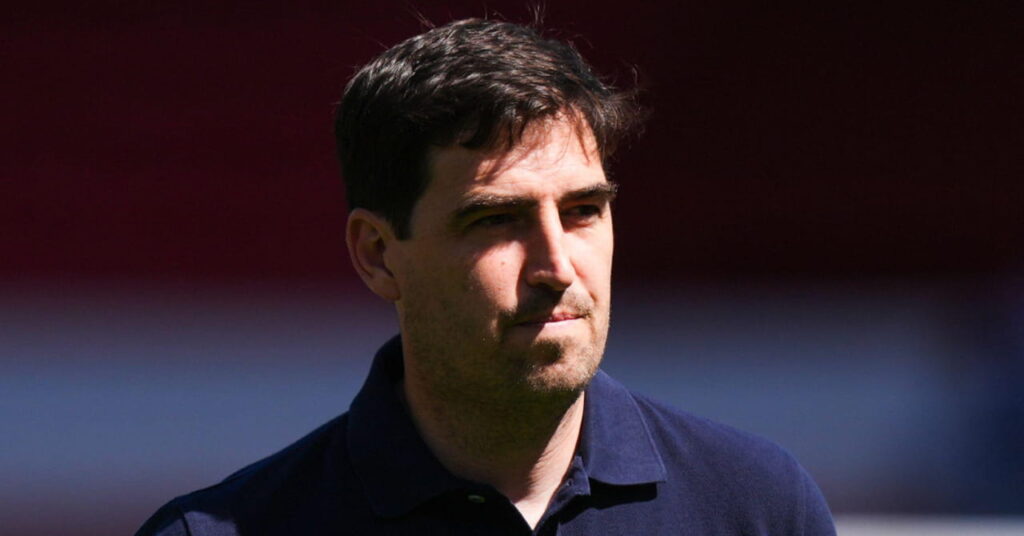 Football manager Andoni Iraola looking to his left while pursing his lips in an outdoor setting with sunshine across his face. The coach is wearing a dark polo shirt