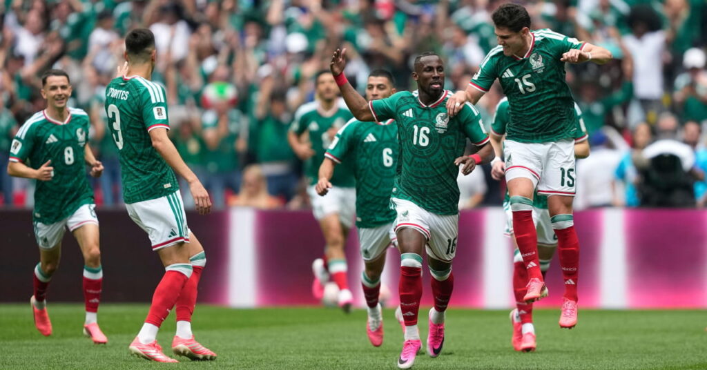 Os jogadores de futebol Israel Reyes e Julian Quinones comemoram um gol com companheiros enquanto jogam pela seleção mexicana de futebol na Copa do Mundo de 2026 e vestem camisas verdes com detalhes brancos, shorts brancos com detalhes vermelhos, meias vermelhas e botas salmão em um campo gramado.