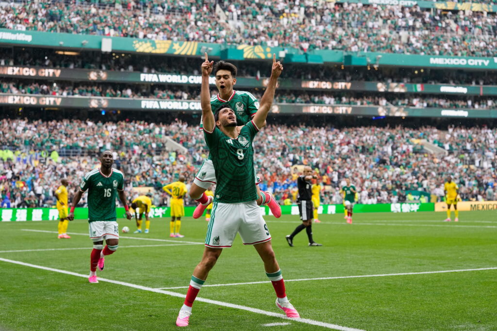O jogador de futebol Raul Jiménez comemora gol com companheiros enquanto joga pela seleção mexicana de futebol na Copa do Mundo de 2026 e veste camisa verde com detalhes brancos, short branco com detalhes vermelhos, meias vermelhas e botas salmão em campo gramado.