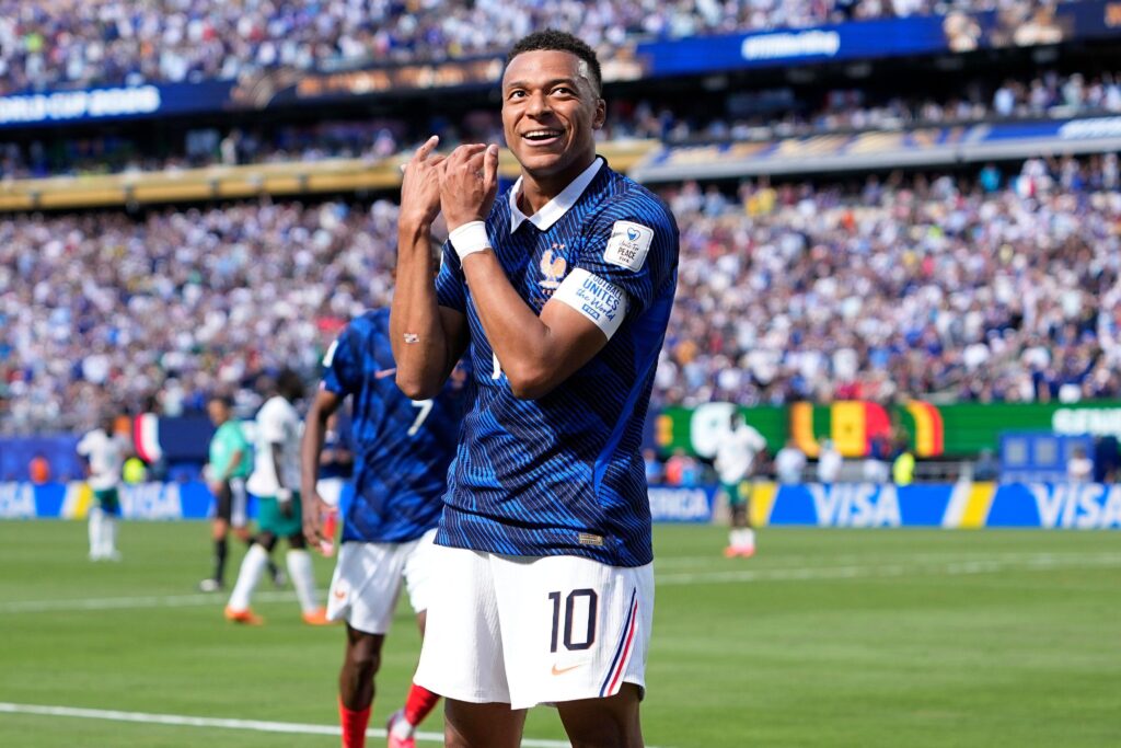 France footballer Kylian Mbappe playing for his national football team in a blue shirt while holding his hands towards his face and smiling after scoring a goal