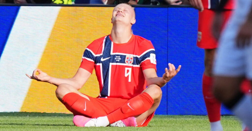 O jogador de futebol norueguês Erling Haaland jogando pela seleção nacional de futebol com uma camisa vermelha e azul com seu número 9 no peito branco e sentado de pernas cruzadas em uma pose de ioga