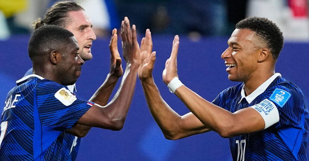 France footballers Ousmane Dembele and Kylian Mbappe exchanging high fives with both hands while on a football pitch during the FIFA football World Cup 2026 and smiling at each other in blue national team shirts, with Adrien Rabiot in the background