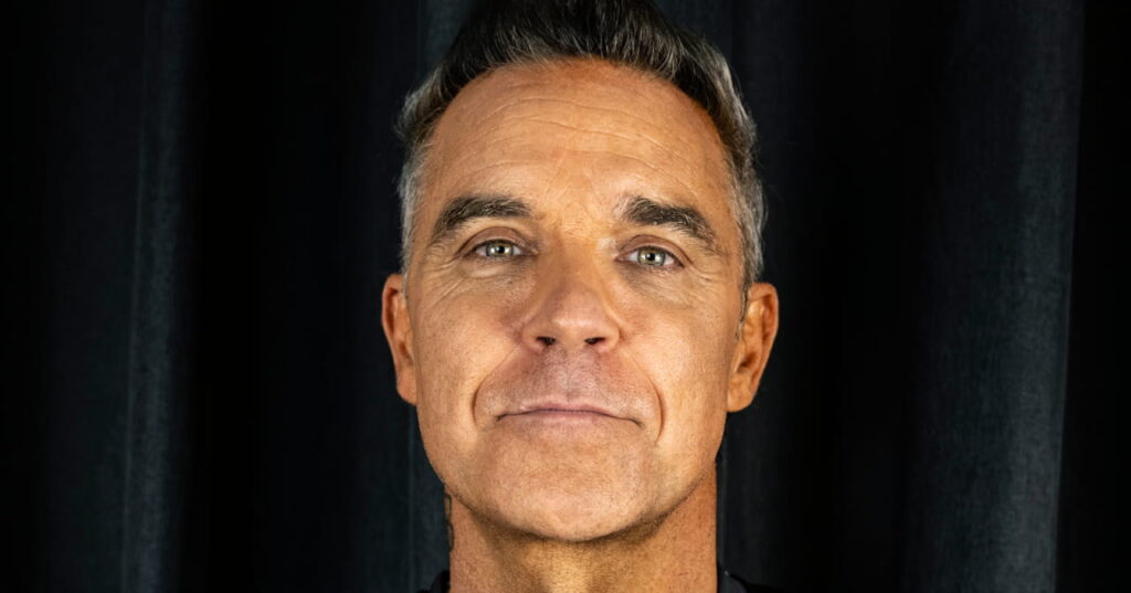 A close up photo of musician Robbie Williams looking forward and smiling at the camera in front of a dark backdrop