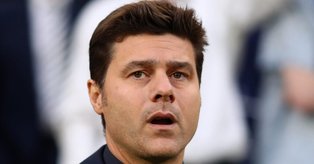 A photo of football manager Mauricio Pochettino looking upwards with an open mouth in a stadium with red seats. The USA head coach is wearing a black coat