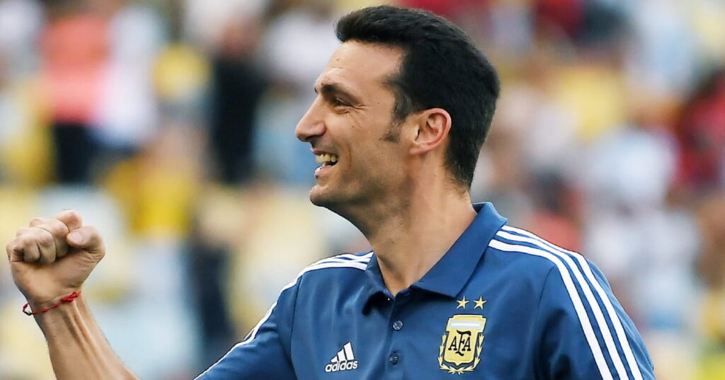 A photo of football manager Lionel Scaloni raising his right fist in a stadium while smiling and celebrating. The Argentina head coach is wearing a blue national team polo shirt with white stripes down the shoulders and a white Adidas logo and yellow national badge on his chest