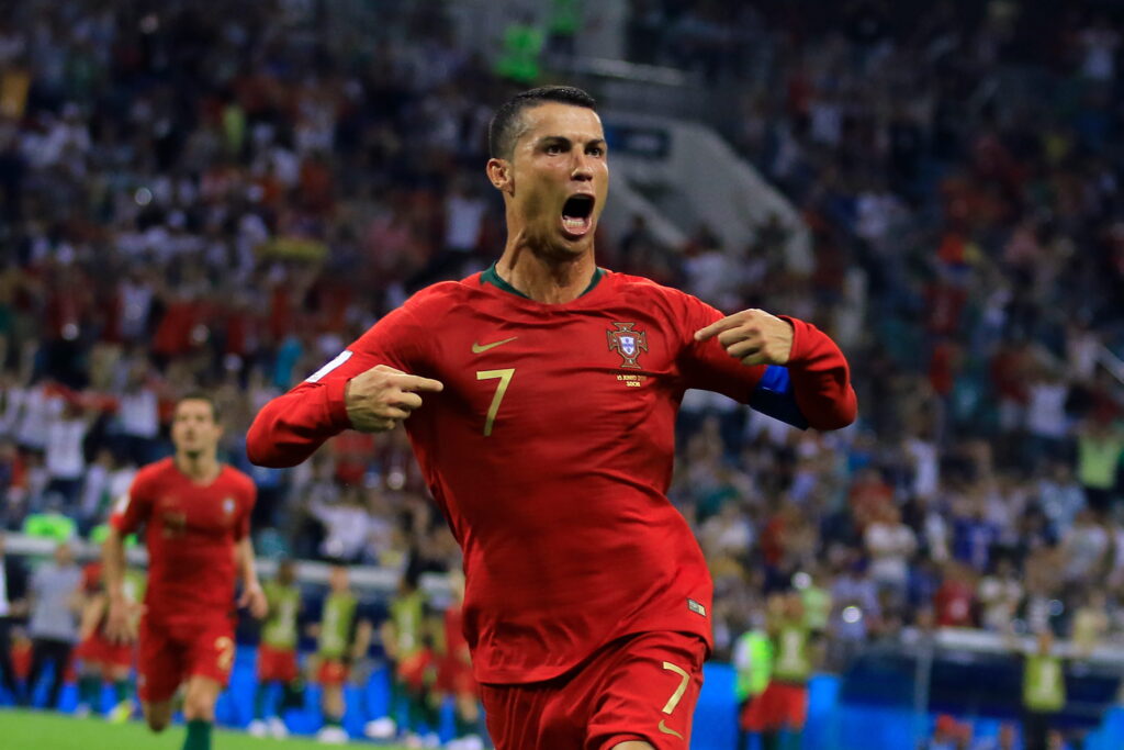 Imagem do jogador de futebol Cristiano Ronaldo comemorando usando as duas mãos para apontar para o peito no campo de futebol. Ronaldo usa o uniforme da seleção portuguesa de futebol com detalhes verdes e pretos e um logotipo e número da Nike branco no peito. Luzes, uma multidão e um companheiro de equipe são levemente visíveis no fundo do estádio